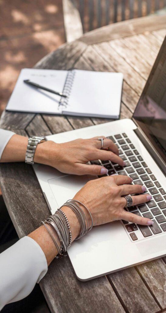 A woman wearing bracelets is typing on a laptop with a notebook and pen in the background