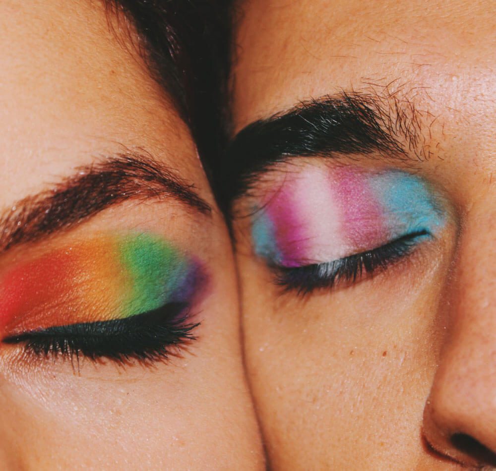 Close-up of two people with rainbow eyeshadow, eyes closed, and eyelashes touching each other's forehead