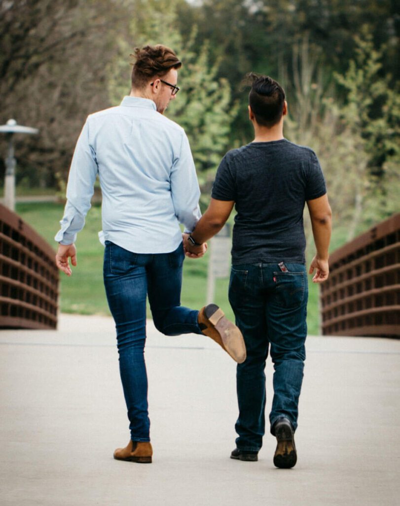 Two young men holding hands and walking across a bridge in a park, one playfully kicks the other