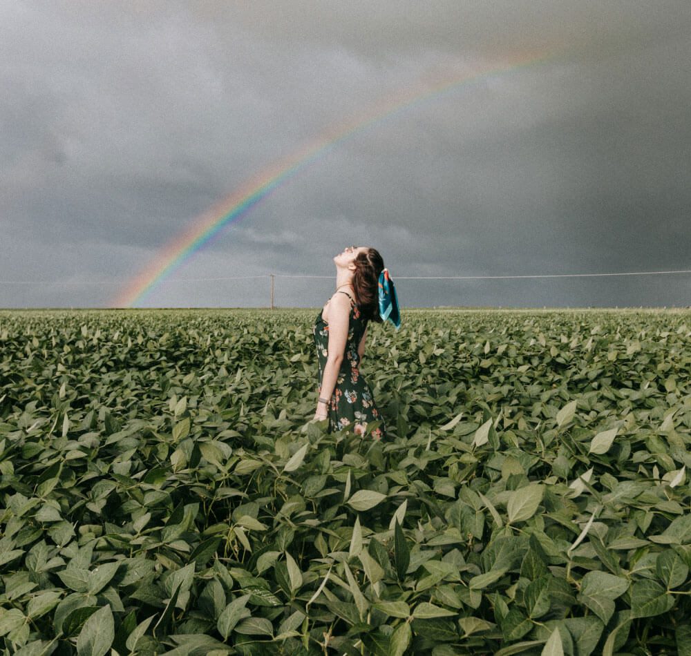 A woman stands in a field with a rainbow in the background