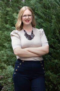 Sarah Perl - A blond woman wearing glasses and a necklace stands with her arms crossed in front of a tree