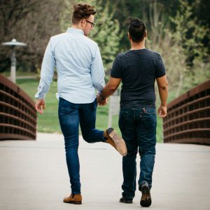 Two young men holding hands and walking across a bridge in a park, one playfully kicks the other
