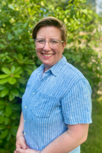 Christa Price stands in front of a bright green wooded area. She is wearing glasses and a blue shirt and has short, blonde-brown hair. She is smiling.