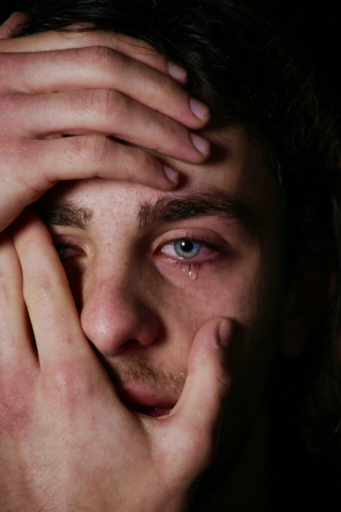 A close up of a young man in a dark space looking at the camera, with his hands partially obscuring most of his face, except for his left eye, which is wet and puffy with a single teardrop lingering on the lower lid.