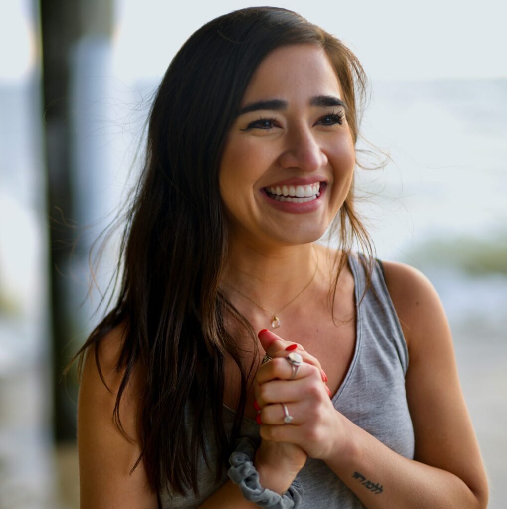 A young woman in a blue blouse stands outdoors. Her hands are clasped together and she smiles broadly while looking off in the distance.
